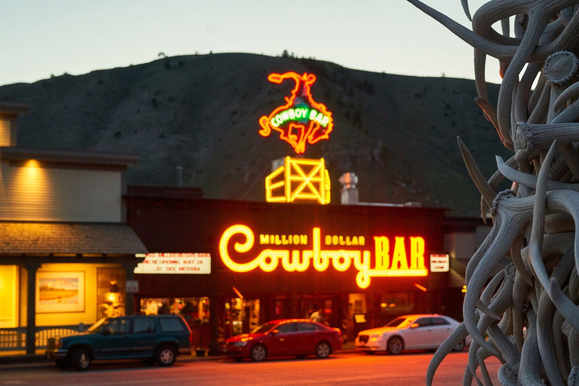 The neon sign of the Million Dollar Cowboy Bar at dusk on the Jackson Town Square, seen past the elk-antler arch